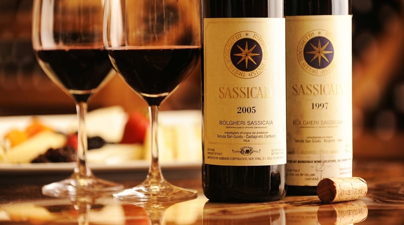 Two glasses of red wine and two bottles of Sassicaia wine with a cork on a reflective surface.