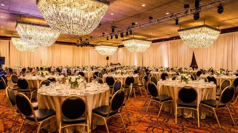A grand ballroom with numerous round tables set for dining, under sparkling chandeliers.