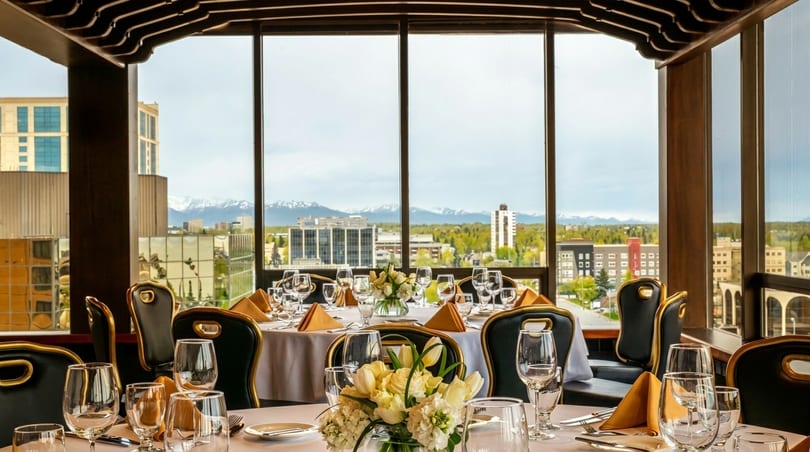 Elegant dining tables with white tablecloths and floral centerpieces, overlooking a city and snowy mountains.