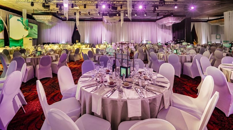 A large ballroom set for an event with numerous round tables, white chairs, and elegant lighting.