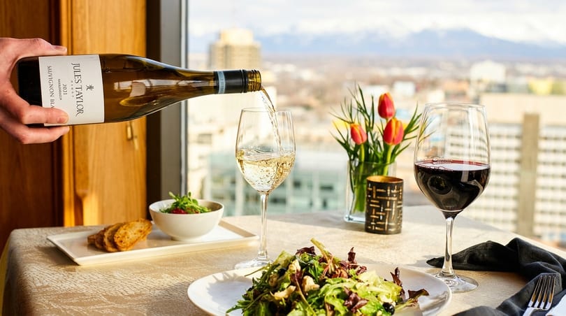White wine being poured into a glass next to a salad, with a city and mountains in the background.